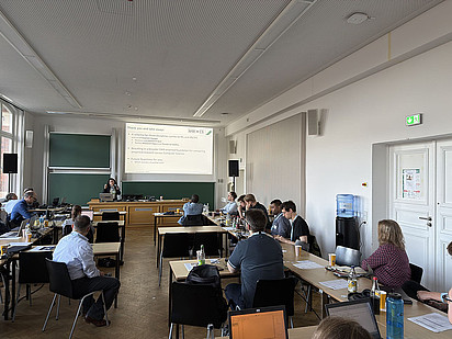 Participants at the 4th NFDIxCS Symposium in Dresden sit at tables in a seminar room, facing a speaker holding a microphone. Behind her, a projected slide titled “Thank you and take-aways” summarises findings on FAIR-oriented empirical research in computer science.