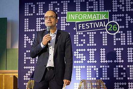 Lars Bernard spricht in ein Mikrofon auf der INFORMATIK26. Hinter ihm ist das Branding der INFORMATIK26 auf einer Wand zu sehen