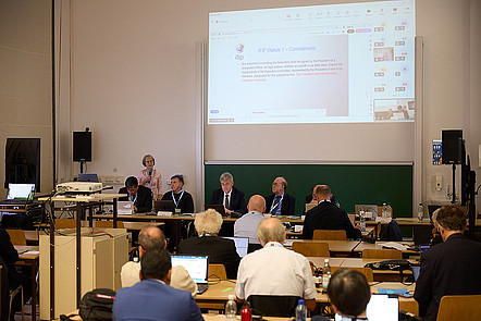 Participants seated at tables with laptops during an IFIP meeting. A woman speaks into a microphone beside the front table, while a projected screen displays “IFIP Statute 7 – Commitments” and remote participants.
