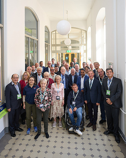 Group portrait of IFIP participants wearing conference badges in a bright corridor at TU Dresden. They face the camera in several rows, with a wheelchair user at the front.